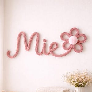 May include: A pink knitted wall decoration spelling "Mie" with a flower accent. The nursery decor also includes a wicker bassinet with a plush blanket and pillow, a stack of towels, and a wooden dresser.