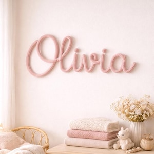 May include: A wall-mounted decorative sign featuring the name Olivia written in a continuous, cursive script. The lettering is crafted from a soft, light pink knitted yarn material, creating a textured, rounded appearance against the wall.