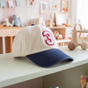 May include: A cream-colored baseball cap with a navy blue brim. The number "3" is embroidered on the front in red with a white outline. The cap is sitting on a light green surface.