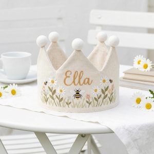Könnte beinhalten: Eine cremefarbene Krone mit dem Namen "Ella" in Gold gestickt. Die Krone hat ein Blumendesign mit Gänseblümchen und Bienen. Weiße Pompons zieren die Spitzen. Die Krone liegt auf einem weißen Tisch.