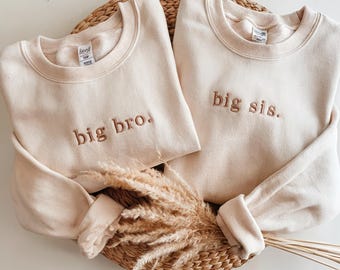 Może przedstawiać: Dwie kremowe bluzy z haftowanym tekstem. Jedna bluza ma napis "big bro.", a druga "big sis."