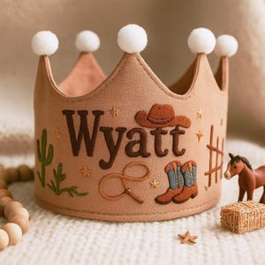 May include: A tan fabric crown with white pom-poms, embroidered with the name "Wyatt", a cowboy hat, cactus, rope, boots, and stars. The crown is a western-themed baby accessory.