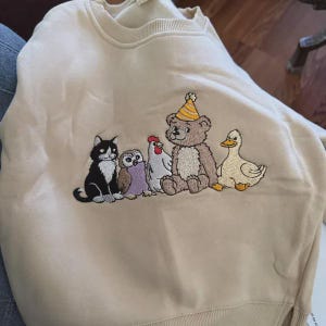 Peut inclure: Un sweat-shirt beige &agrave; col rond orn&eacute; d'une broderie repr&eacute;sentant un chat noir et blanc, un hibou marron, une poule blanche, un ours en peluche marron portant un chapeau de f&ecirc;te ray&eacute; jaune et blanc, et un canard jaune assis en rang.