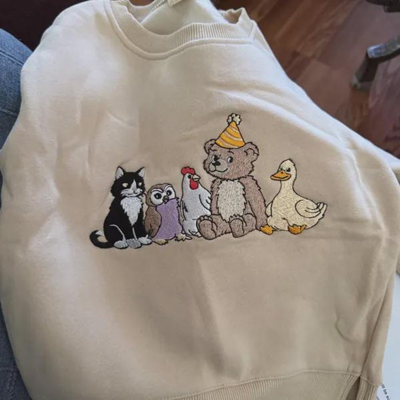 Peut inclure: Un sweat-shirt beige &agrave; col rond orn&eacute; d'une broderie repr&eacute;sentant un chat noir et blanc, un hibou marron, une poule blanche, un ours en peluche marron portant un chapeau de f&ecirc;te ray&eacute; jaune et blanc, et un canard jaune assis en rang.
