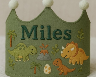 May include: A green fabric crown with the name "Miles" embroidered in dark green. The crown features embroidered dinosaurs, a volcano, and palm trees. Three white pom-poms are attached to the top of the crown.