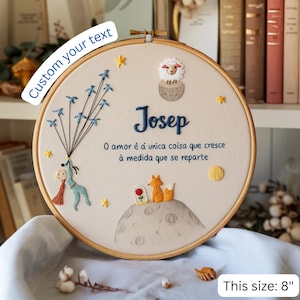 May include: An 20 cm embroidered hoop art piece with a cream-colored background. It features a blue name, Josep, and a Portuguese quote. The design includes a character with birds, a sheep, foxes, and stars.