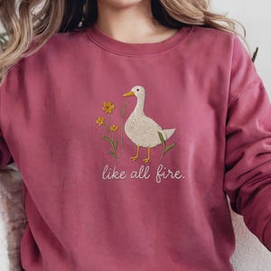 May include: A pink sweatshirt with an embroidered white duck and yellow flowers. The text "like all fire." is embroidered below the duck.