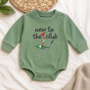 May include: Sage green baby bodysuit with long sleeves. The front features an embroidered golf scene with a red flag, golf ball, and club, along with the text "new to the club" in black script. The bodysuit has snap closures.