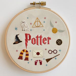 Könnte beinhalten: Gesticktes Bild im Holzrahmen mit dem Wort "Potter" in Rot und thematischen Symbolen. Enthält einen schwarzen Zaubererhut, eine Eule, einen goldenen Schnatz, eine Brille und einen Schal. Das Design ist auf cremefarbenem Stoff.