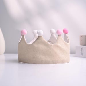 May include: A beige fabric crown with a scalloped edge, topped with alternating white and pink pom-poms. The crown is designed for a child and sits on a white surface. The background is softly lit.