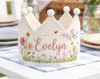 Könnte beinhalten: Eine cremefarbene Krone mit gestickten Blumen, Gras und dem Namen "Evelyn" in rosa Schrift. Die Krone hat weiße Pompons an den Spitzen und eine kleine gestickte Biene. Sie liegt auf einer blau-weiß karierten Oberfläche.