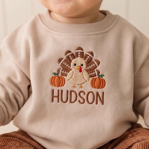 Peut inclure: Un sweat-shirt beige à col rond avec une broderie représentant une dinde aux plumes en forme de ballon de football américain, entourée de deux citrouilles orange. Le prénom HUDSON est brodé en lettres capitales marron sous le motif.
