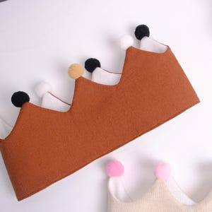 May include: Brown fabric crown with white and black pom-poms. The crown has a zig-zag design with a white interior. A second crown with pink pom-poms is visible in the bottom right corner. The crowns are likely for a child's birthday or costume.