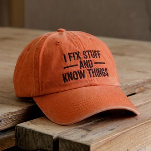 May include: Orange baseball cap with the text "I FIX STUFF AND KNOW THINGS" in black. The cap is made of a soft material and has a curved brim. The hat is sitting on a wooden surface.