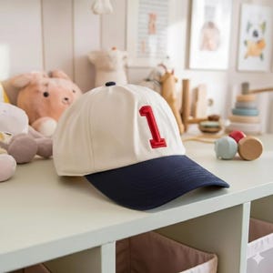 May include: A white and navy blue baseball cap with a red number 1 embroidered on the front. The cap has a curved brim and a small navy blue button on top. The hat is likely for a child.