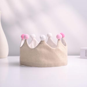 May include: A beige fabric crown with a white interior and adorned with pink and white pom-poms. The crown has a classic shape, suitable for a child's birthday or costume. The crown is sitting on a white surface.