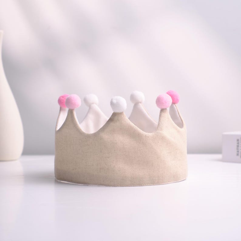 May include: A beige fabric crown with a white interior and adorned with pink and white pom-poms. The crown has a classic shape, suitable for a child's birthday or costume. The crown is sitting on a white surface.