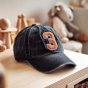 May include: A dark gray baseball cap with a curved brim. The cap features a large orange number "3" patch outlined in white on the front. The cap is sitting on a light wood surface.