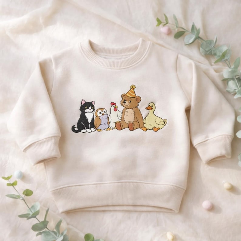 Peut inclure: Un sweat-shirt &agrave; manches longues couleur cr&egrave;me avec une impression graphique repr&eacute;sentant un chat noir et blanc, un hibou brun, une poule blanche, un ours en peluche brun portant un chapeau de f&ecirc;te jaune et un canard jaune align&eacute;s.