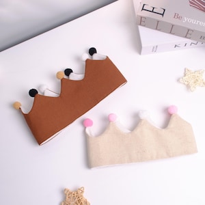 May include: Two fabric crowns with pom-pom details. One crown is brown with black, white, and tan pom-poms. The other crown is beige with pink and white pom-poms. The crowns are on a white surface.