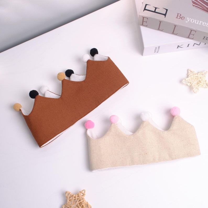 May include: Two fabric crowns with pom-pom details. One crown is brown with black, white, and tan pom-poms. The other crown is beige with pink and white pom-poms. The crowns are on a white surface.