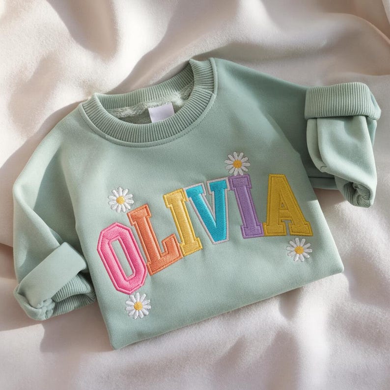 May include: A pale green sweatshirt featuring the name "OLIVIA" in embroidered, multi-coloured letters. The letters are outlined in a contrasting colour. Small daisy flower designs are embroidered around the name. The sleeves are cuffed.