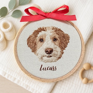 May include: A circular embroidered dog portrait in a wooden hoop, depicting a brown and white dog with the name "Lucass" stitched below. A red satin bow is tied at the top. The image is on white fabric, with baby booties and a wooden toy.
