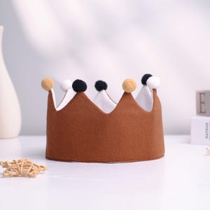May include: A brown fabric crown with a white interior and topped with alternating pom-poms in tan, white, and black. The crown is sitting on a white surface. The crown is a festive accessory.