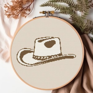Könnte beinhalten: Ein brauner Cowboyhut mit weißem Rand und braunem Band ist auf eine beige Leinwand gestickt.