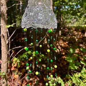 May include: A clear, cut-glass wind chime with a decorative pattern hangs from a black cord. Strands of green and yellow beads dangle below the glass, creating a colorful and whimsical outdoor decoration. The background features a natural, wooded setting.