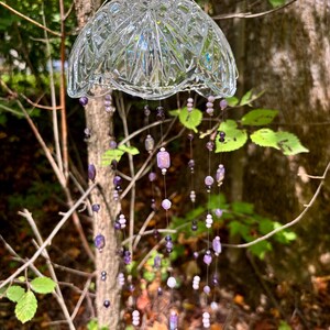 May include: A clear glass wind chime with a decorative, patterned bowl shape. Strands of purple and white beads hang below. The wind chime is suspended from a tree branch, creating a whimsical outdoor decoration.