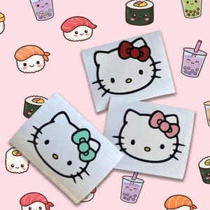 May include: Three white Hello Kitty stickers with different colored bows: red, pink, and teal. The stickers are surrounded by cartoon sushi and bubble tea illustrations on a pink background.