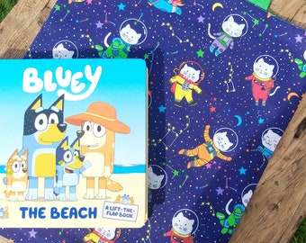 Può includere: Una borsa per libri blu con un manico verde e un tessuto blu scuro con un motivo di gatto astronauta bianco. La borsa è piena di un libro blu e giallo intitolato "Bluey The Beach A Lift-The-Flap Book."