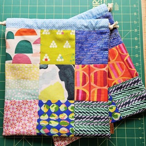 Puede incluir: Una bolsa con cordón de patchwork con una variedad de cuadrados de tela coloridos. Los cuadrados presentan patrones abstractos en tonos de rosa, verde, amarillo, azul y naranja. La parte superior de la bolsa es azul claro con cierre de cordón.