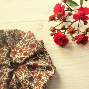以下が含まれることがあります： 赤と青の花柄の白い布製のシュシュ。シュシュは明るい木製の表面に赤いバラの枝と一緒に置かれています。