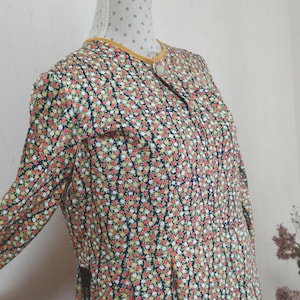 May include: A long-sleeved dress with a floral print in shades of red, yellow, white, and green on a dark blue background. The dress has a yellow trim around the neckline and a button closure at the front.