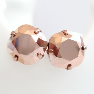 May include: A pair of stud earrings featuring square-cut, rose gold-toned crystals with faceted surfaces. Each crystal is securely held in place by four small, matching rose gold-toned metal prongs, creating a clean and reflective appearance.
