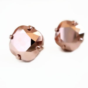 May include: A pair of stud earrings featuring square-cut, faceted rose gold crystals. Each crystal is secured in a four-prong metal setting that matches the metallic rose gold tone of the stone, creating a cohesive and reflective appearance.