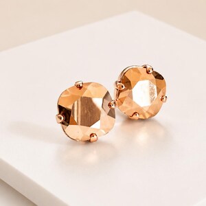 May include: A pair of stud earrings featuring square-cut, faceted champagne-colored crystals set in rose gold-toned metal prongs. The earrings have a reflective, polished surface and a symmetrical design, showcasing a warm metallic hue throughout.