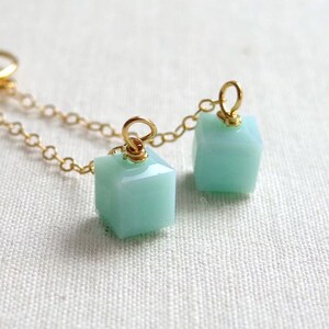 May include: Two light mint green cube-shaped beads attached to a thin gold-colored metal chain. Each bead features a faceted surface and is secured to the chain by a small gold-colored metal jump ring wrapped around the top of the cube.