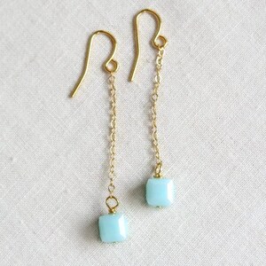 May include: A pair of gold-tone drop earrings featuring a delicate chain suspended from a curved ear wire. Each earring terminates in a single, small, square-shaped light blue bead with faceted edges, creating a simple and clean design.