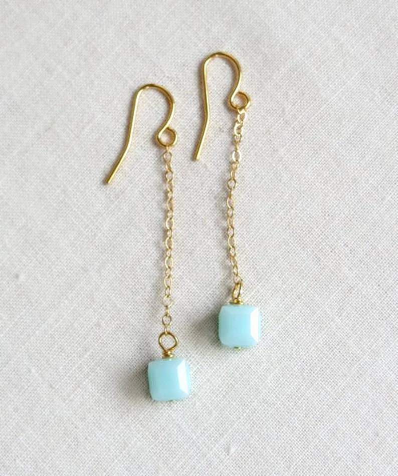 May include: A pair of gold-tone drop earrings featuring a delicate chain suspended from a curved ear wire. Each earring terminates in a single, small, square-shaped light blue bead with faceted edges, creating a simple and clean design.