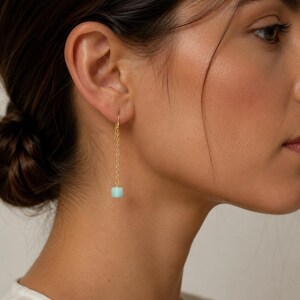May include: A gold-tone drop earring featuring a delicate chain suspended from a hook, ending in a single cylindrical light blue bead. The bead has a diameter of 6mm. The earring hangs vertically, showcasing a minimalist design with a polished metal finish and a smooth, matte-textured stone component.