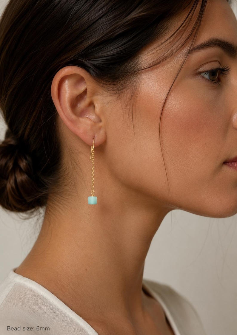 May include: A gold-tone drop earring featuring a delicate chain suspended from a hook, ending in a single cylindrical light blue bead. The bead has a diameter of 6mm. The earring hangs vertically, showcasing a minimalist design with a polished metal finish and a smooth, matte-textured stone component.