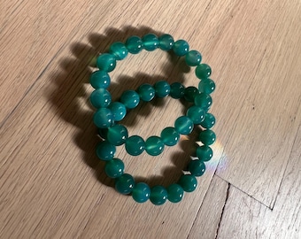 May include: Two jade green beaded bracelets. Each bracelet is made of round, polished beads. The bracelets are stacked on top of each other, creating a layered effect. The beads have a glossy finish and reflect light.