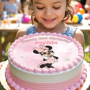 May include: A two-tiered pink and white cake decorated with a Minnie Mouse image and the words "Happy 2nd Birthday Sophia". The cake is held by a young girl. The background shows a garden party with other children and decorations.