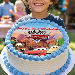 May include: A birthday cake with blue frosting and a Cars-themed image. The image features the text "Happy 2nd Birthday" and the name "Thomas". The cake is held by a smiling child.