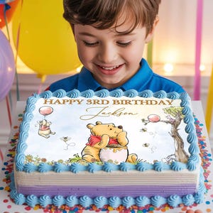 Op de afbeelding: Een kind houdt een rechthoekige verjaardagstaart vast, versierd met een Winnie de Poeh-thema. De taart heeft blauwe glazuur, een paarse streep en de woorden "Happy 3rd Birthday Jackson". Winnie de Poeh wordt afgebeeld met een honingpot.