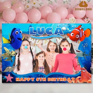 May include: A blue and orange photo booth backdrop with a Finding Nemo theme. The backdrop features the characters Dory, Nemo, and a shark. The backdrop also has a space for a photo with the text "Luca" and "Happy 5th Birthday".