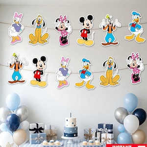 Pode incluir: Um conjunto de decoração de festa com duas guirlandas de personagens da Disney recortados, incluindo Mickey Mouse, Minnie Mouse, Pato Donald, Margarida, Pateta e Pluto, exibidos sobre uma mesa com um bolo de dois andares, presentes e balões azuis e prateados.